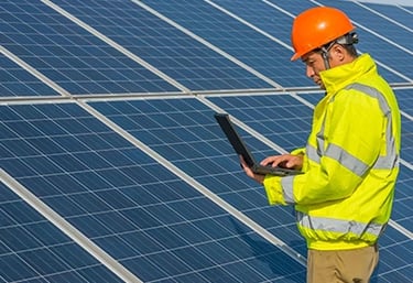 Solar energy technician in a hard hat using a laptop to inspect a solar panel farm.