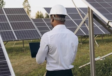 Solar energy engineer in a white hard hat using a laptop to inspect a solar panel field.