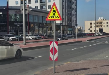 Yellow pedestrian crossing road sign on a city street with cars and urban buildings.