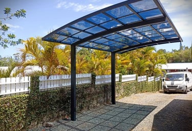 Modern black metal carport with blue tinted glass panels over a gravel driveway next to palm trees.