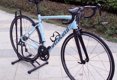Light blue Specialized Allez road bike on a display stand with carbon fiber forks and drop handlebars.