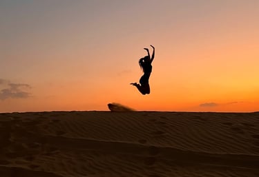 An image of a lady jumping over the sand dunes with a beautiful sunset on the background