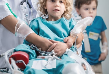 A caregiver wearing surgical attire reads 'The Cat in the Hat' to a child in a protective environment. The child, also wearing protective clothing, smiles while clutching a doll. The setting appears to be a medical isolation room, indicated by the plastic curtains and sterile equipment.