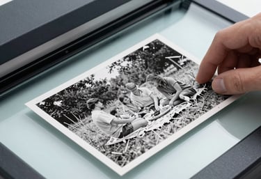 Close-up photography of a weathered, torn black and white photo of a family picnic in a North American / US setting. A hand is placing it onto the glass of a professional scanner. Clean, modern aesthetic with Slate Blue tones.
