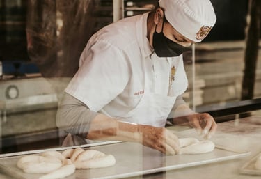 Panadero profesional con uniforme blanco y cubrebocas dando forma a masa fresca sobre una bandeja pa