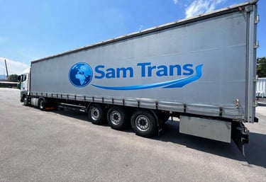 A silver Sam Trans logistics semi-truck trailer parked on asphalt under a clear blue sky.