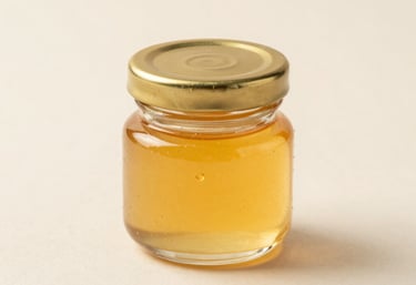 Professional product photography of a small 100ml clear glass jar with a golden lid, showcasing viscous honey yellow liquid, placed on a clean cream surface.