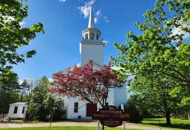 The historic East Winthrop Baptist Church with white siding, a steeple, and a flowering pink tree.