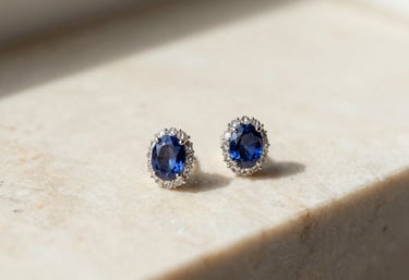 An elegant pair of sapphire and diamond earrings resting on a warm cream marble surface. The composition is clean and airy with soft, natural morning light.