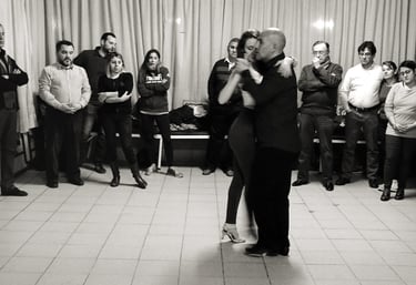 Una pareja bailando tango argentino en un estudio de baile, con alumnos observando.