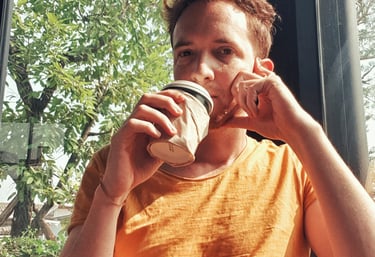 Unicris with a geometric arm tattoo drinking coffee by a sunlit cafe window.