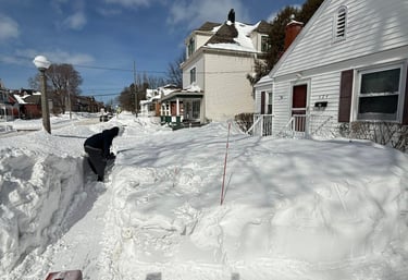 shoveling a deep snow drift on a sidewalk near a snowblower.