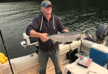 A smiling fisherman holds a freshly caught silver salmon on a boat in calm coastal waters.