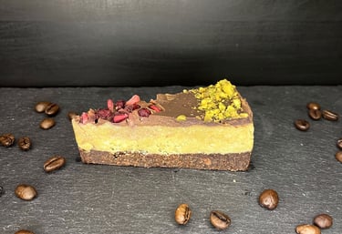 A slice of vegan raw cake with pistachio and dried berries on a slate board with roasted coffee beans.