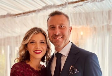 A smiling couple dressed in formal wedding guest attire posing at an elegant indoor event venue.