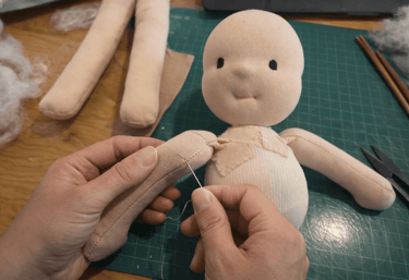 Manos cosiendo brazos de una muñeca y al fondo se ve la cabeza y parte del pecho de una muñeca waldorf en proceso