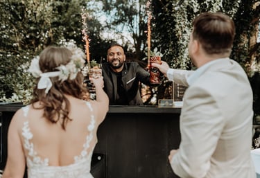 A smiling wedding bartender serves sparkling cocktails to a bride and groom at an outdoor garden bar.
