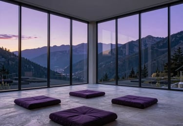 Architectural photography of a minimalist meditation hall in the North American mountains. Large glass walls reveal a purple-hued sunset. The interior features light grey stone and dark purple cushions. Clean, sophisticated style.