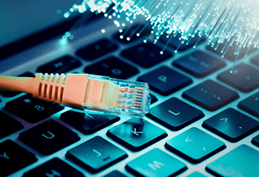 An Ethernet cable connector resting on a laptop keyboard next to glowing blue fiber optic cables.