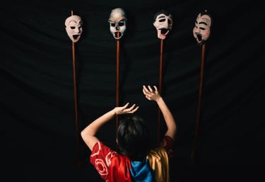 A theatrical performer reaches toward four expressive hand-painted theater masks on wooden poles.