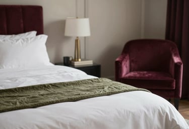 A cozy, luxuriously appointed bedroom with high-thread-count parchment linens, a smoky olive throw, and a deep burgundy velvet armchair in the corner. Elegant and inviting. Palette includes #FBF9F4, #6F7A6E, and #7D2F45.