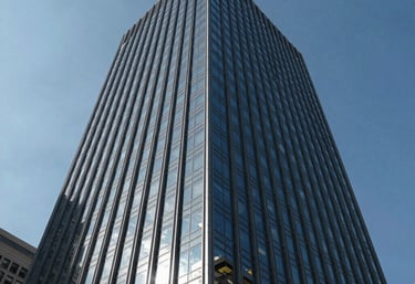 A low-angle exterior shot of a modern glass skyscraper in a North American / US city reflecting the blue sky. The image represents stability and commercial success.