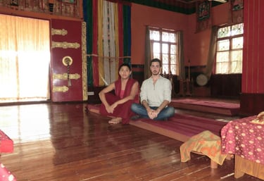 A Buddhist monk and a traveler sit cross-legged on mats inside a traditional Tibetan temple sanctuary.