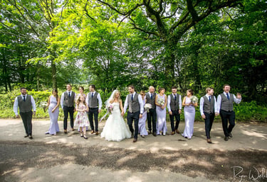 A bride and groom walk with their wedding party down a tree-lined path during a summer woodland wedding.