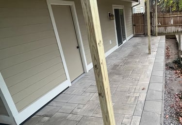 Custom stone paver patio installed under a raised wooden deck with tan vinyl siding.