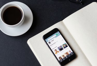 A top-down view of a International / Global professional's workspace with a coffee cup, a notebook, and a smartphone showing a business app. The colors are dark navy and soft off-white.