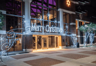 A brick building entrance decorated with a glowing white Merry Christmas neon sign and festive holiday lights.