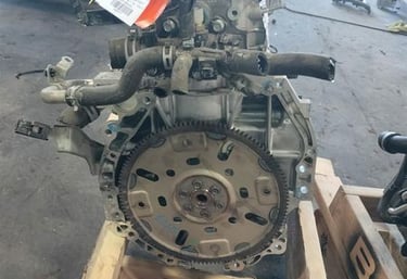 Used car engine assembly with flywheel on a wooden pallet in an auto parts warehouse.