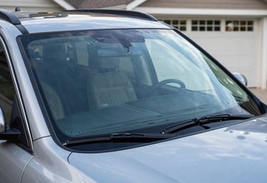 A photograph showing the finished result of a windshield replacement on a luxury SUV. The vehicle is parked in a clean North American driveway, reflecting the professional steel blue sky and house architecture in the flawless new glass.