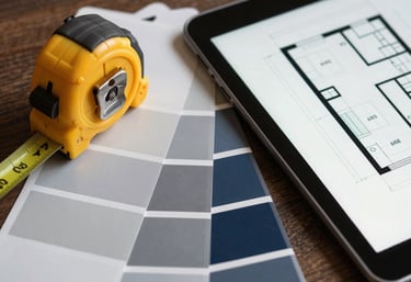 A stylized flat-lay image of modern interior design tools including a measuring tape, color swatches in gray and navy, and a digital tablet showing a floor plan.