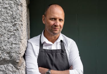 Professional chef wearing a white shirt and black apron leaning against a stone wall.