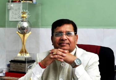 Professional man in white Nehru jacket sitting at a desk with an Indian flag and trophies.
