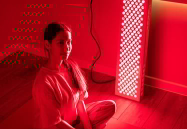 A woman sits in front of a red light therapy panel for skin health and recovery.