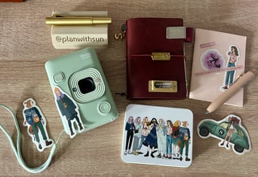 Flat lay of an Instax Mini LiPlay camera, leather journal, and journaling stickers on a wooden desk.