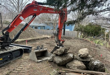 Orange Kubota excavator moving large boulders for a backyard landscape retaining wall project.