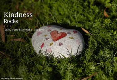 Hand-painted kindness rock with a red heart sitting in green grass, promoting spreading love and positivity.