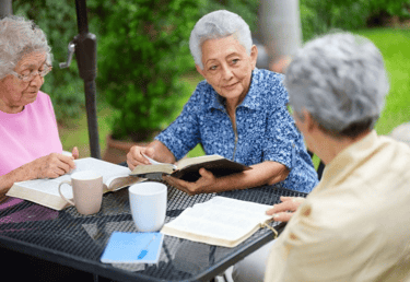 Three elderly women engaged in a book club meeting outdoors, exemplifying senior participation in li