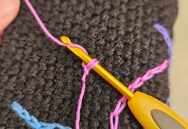 A yellow crochet hook making pink surface slip stitches on a dark brown textured fabric.