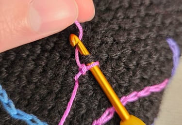 Close-up of a hand using a gold crochet hook to surface crochet pink yarn onto black fabric.