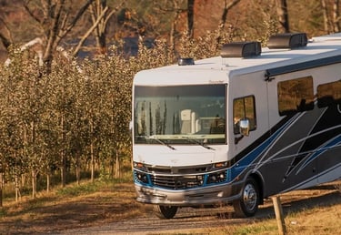 A white Fleetwood Bounder Class A motorhome driving through a scenic rural apple orchard at sunset.