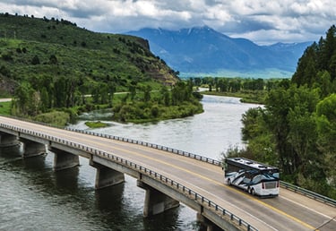 A luxury motorhome drives across a bridge over a scenic river with mountain views.