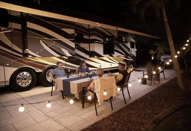 Friends enjoy outdoor dining by a luxury motorhome RV at night with festive string lights.