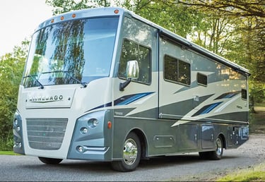 A gray and tan Winnebago Vista Class A motorhome parked on a paved road surrounded by lush green trees.