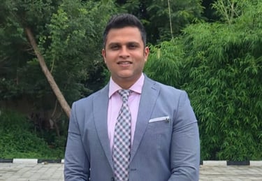 Professional businessman in a grey suit and pink shirt standing outdoors with a green tree background.