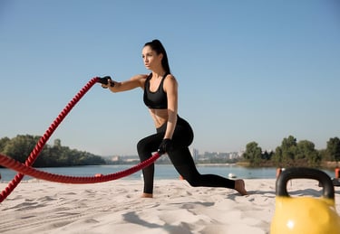 Mulher treinando com corda naval na areia, próximo a um lago, em dia ensolarado.