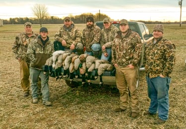 Six hunters in camouflage gear standing in a field behind a pickup truck loaded with harvested ducks and geese.